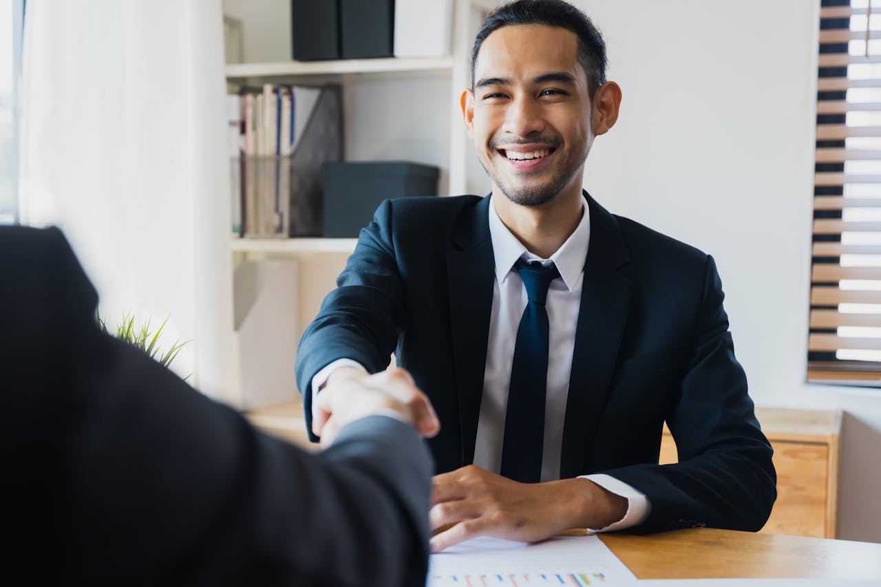 hero-img-02 Confident businessman in suit shaking hands at office desk, symbolizing successful partnership.